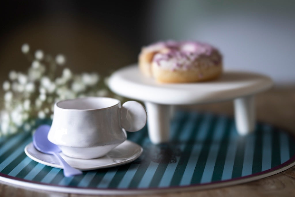 White cup and saucer on a striped table with a donut on a stand in the background.